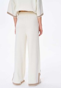 Wijd-uitlopende crèmewitte broek met lichtbruine afwerking. Zacht, gestructureerd materiaal; heeft zijslits aan de zoom voor extra detail. Elastische tailleband.