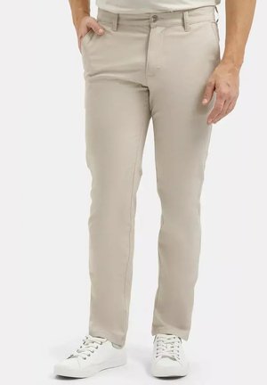 Mężczyzna ubrany w beżowe wąskie spodnie i białe trampki, z rękami spoczywającymi po bokach na białym tle.