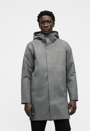 Homme portant une longue veste imperméable grise avec capuche et poignets pression, debout devant un fond clair uni.