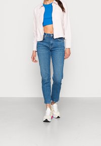 Ljusrosa kort jacka över en blå kort topp, matchad med medelhöga blå jeans och vita sneakers med rosa detaljer.