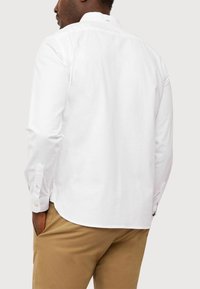 Camicia bianca a maniche lunghe con colletto a band, caratterizzata da polsini con bottoni e una texture liscia. Cuciture visibili e spacchetti sui lati dell'orlo.