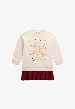 Haut à manches longues couleur crème avec logo Guess en paillettes dorées, petits accents cœur rouge et étoile, et détail d'ourlet de jupe plissée bordeaux.