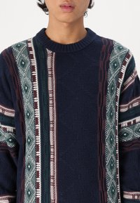 Námořnický pletený svetr s propracovanými vzory v zelené, bordó a bílé barvě, s žebrovaným kulatým výstřihem a texturovanou tkaninou.