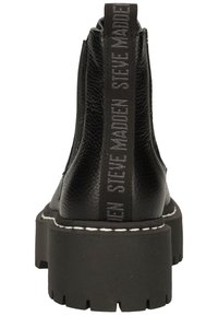Schwarze Knöchelstiefel aus strukturiertem Leder, mit einer klobigen Plattformsohle und einem hochgezogenen Rückenteil, das "STEVE MADDEN" in Grau zeigt. Weiße Nähte.