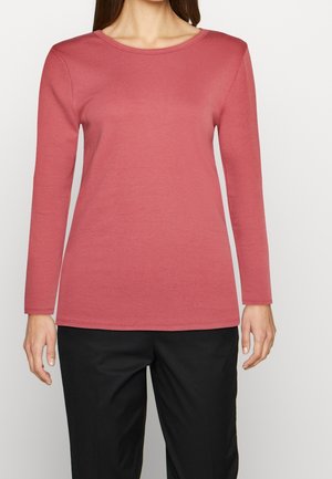 Woman wearing a long-sleeve, crew neck, dusty pink shirt paired with black trousers, standing against a plain light background.