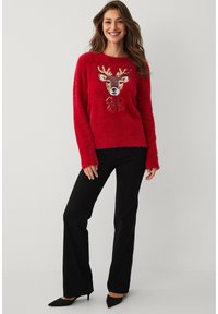 Rode zachte trui met een ontwerp van een hertenhoofd en details van pailletten, gecombineerd met zwarte uitlopende broek en puntige zwarte schoenen.