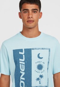 Lyseblå kortærmet t-shirt lavet af blødt stof, med et blåt grafisk design, der indeholder elementer som sol, måne og palmetræ.