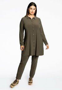 Olijfgroene tuniek met een knoopsluiting aan de voorkant, lange mouwen en een rechte snit, gecombineerd met bijpassende aangesloten broek en beige sneakers.