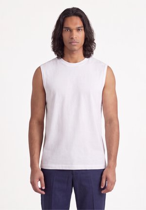Uomo con capelli scuri fino alle spalle, che indossa una camicia bianca senza maniche e pantaloni blu scuro, in piedi davanti a uno sfondo chiaro uniforme.