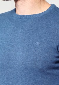 Pull en maille bleu avec un col rond et un petit logo sur la poitrine. Le tissu semble lisse et légèrement texturé.