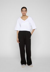 Mujer de pie mirando al frente, vestida con una blusa blanca de cuello en V y mangas hasta el codo, pantalones negros sueltos y zapatillas blancas, con la mano en el bolsillo.