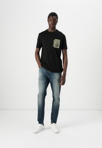 Un giovane uomo si trova di fronte a uno sfondo semplice, indossando una maglietta nera con una tasca decorata, jeans chiari e sneakers bianche.