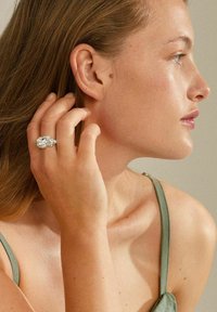 Anillo de plata con una gran piedra preciosa, llevado en una mano que toca el cabello. La persona tiene el cabello castaño claro y viste una blusa verde de satén con tirantes. Fondo neutro.