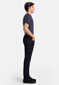 Mörkblå jeans med slim fit, bärs med en marinblå t-shirt. Modellen har händerna i fickorna och har på sig svarta slip-on skor. Enkelt design.
