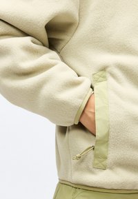 Χέρι στην πλαϊνή τσέπη μπεζ φλις μπουφάν με φερμουάρ στην τσέπη και λεπτομέρεια με κορδόνι, συνδυασμένο με πράσινο παντελόνι.