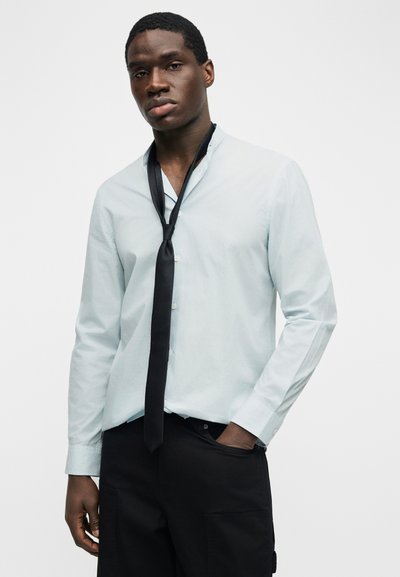 Νεαρός άνδρας που φορά ένα γαλάζιο πουκάμισο με κουμπιά και μια μαύρη γραβάτα που δεν είναι δεμένη, στέκεται με το ένα χέρι στην τσέπη του μαύρου παντελονιού.