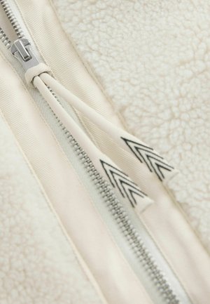 Detalle de chaqueta de forro polar con tela blanca texturizada, cremallera plateada y cinta de cremallera color crema con acentos en chevron negro.