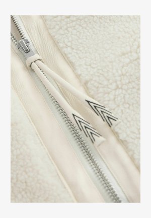 Detailansicht einer Fleecejacke mit einer strukturierten weißen Stoffoberfläche, silbernem Reißverschluss und cremefarbenem Reißverschlussband mit schwarzen Chevron-Akzenten.