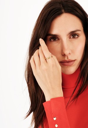 Anello ondulato in oro indossato su un dito, con una finitura liscia. La persona che lo indossa ha i capelli lunghi e scuri ed è vestita con un maglione girocollo rosso.