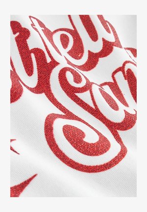 Rote glitzernde Schrift auf weißem Stoff, mit dekorativen, geschwungenen Buchstaben und einer glänzenden, strukturierten Oberfläche. Nahaufnahme hebt die Details hervor.