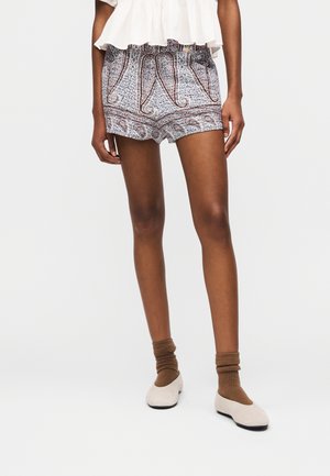 Modello che indossa pantaloncini con motivo paisley, calzini marroni, ballerine beige e una maglietta bianca con volant, in piedi su uno sfondo chiaro e uniforme.