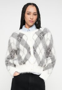 Fehér és szürke színű, mintás argyle mintás bolyhos kardigán, kerek nyakkivágással, hosszú ujjakkal és krémszínű gombokkal. Világoskék ing fölött viselve.