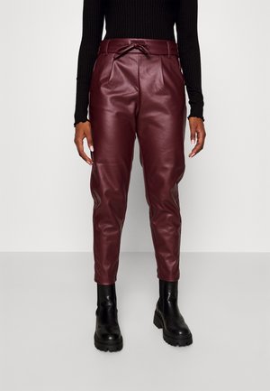 Osoba ubrana w wysokowaistowe burgundowe spodnie ze sztucznej skóry z wiązaniem z przodu, połączone z czarnymi masywnymi botkami i czarną prążkowaną bluzką.