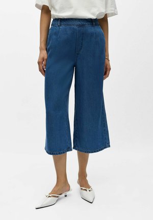 Femeie purtând blugi croiți largi, scurtați, din denim albastru, top alb și pantofi albi cu toc mic ascuțit și detaliu cu fundă.