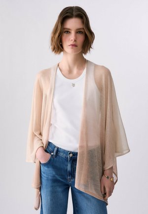 Donna con capelli castani corti che indossa un cardigan beige trasparente sopra una maglietta bianca e jeans blu, una mano in tasca, sfondo neutro.