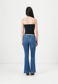 Crna bešavna majica, plave traperice s visokim strukom i širokim nogavicama s dvostrukim stražnjim džepovima, denim tekstura i drveni detalj na pojasu.