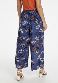 Bloemrijke wijde broek in blauwe stof, met een levendig bloemenpatroon, een elastische tailleband en witte sleehakken.