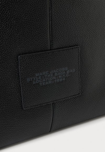 Détail d'un sac en cuir noir texturé avec une étiquette rectangulaire cousue affichant un texte en relief de la marque et du style à peine visible.