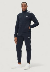 Marinträningsdräkt med zip-up jacka, ribbade ärmslut och logodetaljer. Matchande joggers och vita sneakers. Slät tygkvalitet.
