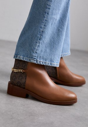 Brune læderankelstøvler med mønstret stofbagstykke og guldkæde-detalje, båret med lyseblå cowboybukser på gråt gulv.