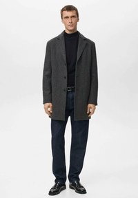 Manteau gris foncé en mélange de laine avec revers crantés, coupe à simple boutonnage, porté sur un col roulé noir. Associé à un jean foncé et des chaussures noires.