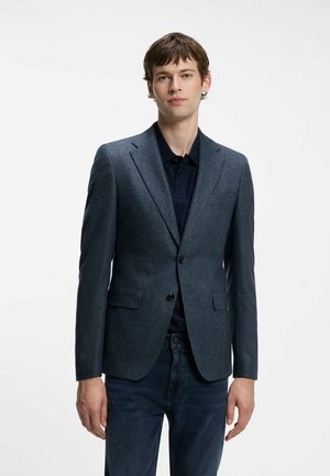 Joven con una blazer azul texturizada sobre una camisa oscura y vaqueros oscuros, de pie frente a un fondo blanco liso.