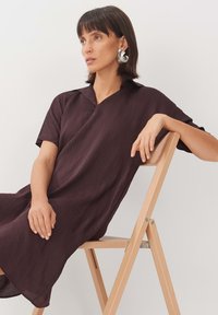 Dunkelburgunderfarbendes Leinenkleid mit Kragen, kurzen Ärmeln und lockerer Passform. Das Model sitzt auf einem Holzstuhl und präsentiert die Struktur des Kleides.