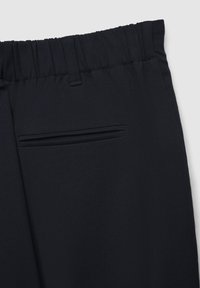 Nahaufnahme von schwarzen Hosen mit elastischem Bund und einer einzelnen Paspeltasche auf glattem Stoff.