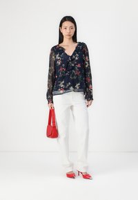 Blouse florale marine avec un col à volants, manches longues transparentes, associée à un pantalon blanc à jambes larges, un sac à main rouge et des escarpins rouges à bout pointu.