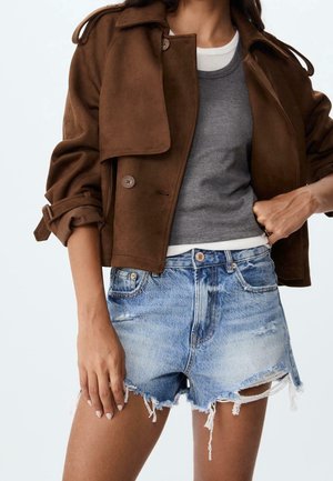 Persoon draagt een bruin jasje over een grijs mouwloos topje en een wit overhemd, gecombineerd met lichtblauwe, hoog getailleerde, versleten spijkershorts.