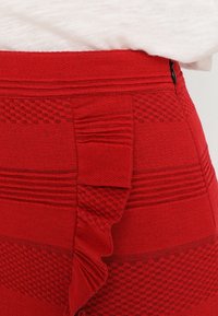 Červená pletená sukně s texturovanými vzory, horizontálními pruhy a volánkovým zdobením na boku. Hladká tkanina s přiléhavým střihem.