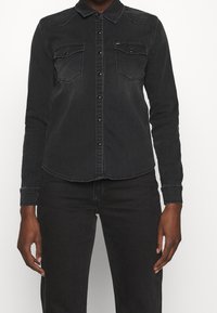 Camicia in denim nero con maniche lunghe, chiusura con bottoni frontale, due tasche sul petto, orlo arrotondato e dettagli di cucitura sottili.