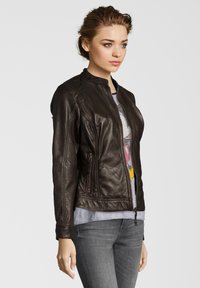 Braune Lederjacke mit taillierter Passform, Reißverschluss vorne und seitlichen Taschen. Rippenakzente an den Schultern und an den Manschetten. Getragen über einem Grafik-T-Shirt.