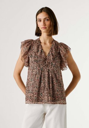 Frau trägt eine gemusterte, ärmellose Bluse mit gerafften Schultern und V-Ausschnitt, kombiniert mit einer weißen Hose, steht mit den Händen in der Hüfte.