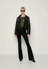 Corta-vento de couro preto sobre uma blusa verde-azeitona, combinado com calças pretas de perna larga e botas pretas de bico fino. Apresenta óculos de sol estreitos.
