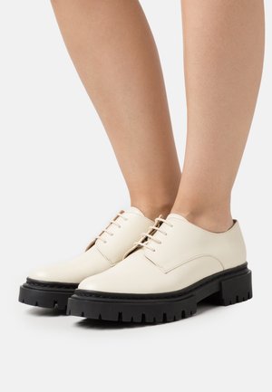 Chaussures en cuir beige avec un bout rond, dotées de cinq œillets et de semelles en caoutchouc noir avec un motif de bande de roulement robuste.