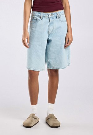 Personne portant un short en jean bleu clair jusqu'aux genoux, des chaussettes blanches, des chaussures beiges sans lacets et un haut sans manches bordeaux.