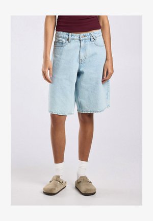 Persona con pantalones cortos de mezclilla azul claro hasta la rodilla, calcetines blancos, zapatos beige sin cordones y una camiseta sin mangas burdeos.