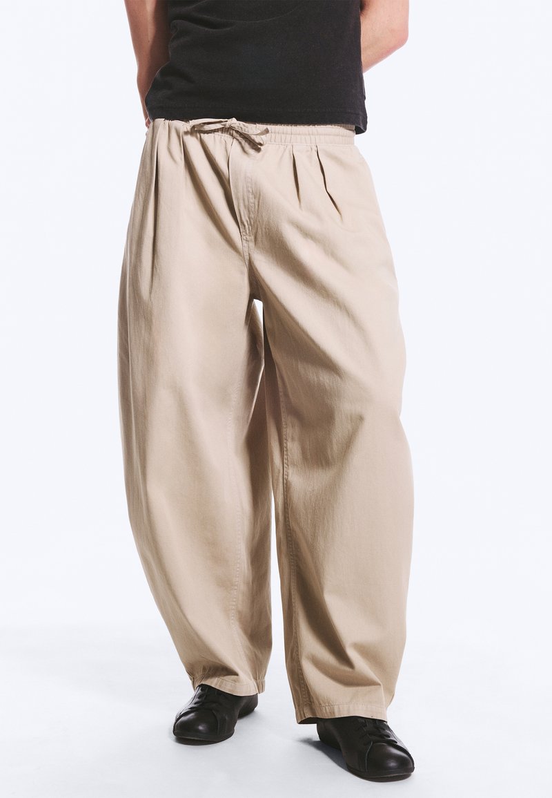 Personne portant un pantalon beige ample avec taille à cordon et chaussures noires, debout sur fond blanc.