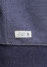 Tela azul marino con un patrón texturizado de puntos; incluye una etiqueta blanca con el texto "HILFIGER" y "NEW YORK NY" en azul.